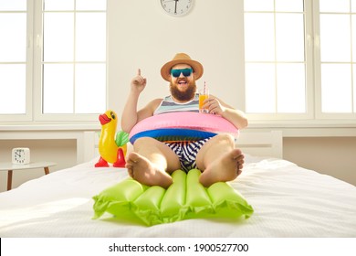 Happy young man spends summer holidays in Covid pandemic quarantine. Chubby male tourist lying on bed with inflatable lifebuoy and beach mattress, drinking juice and enjoying 'stay at home' vacation