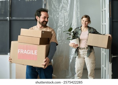 Happy young man with stack of cardboard boxes with fragile household stuff moving along living room with his wife behind