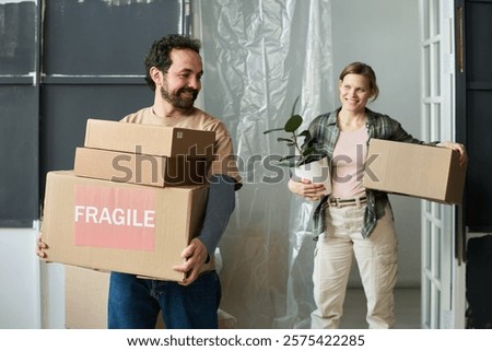 Happy young man with stack of cardboard boxes with fragile household stuff moving along living room with his wife behind