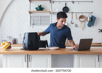 Happy Young Man Standing At Kitchen Table And Cooking Toast Bread By Air Fryer Machine, Watching Culinary TV Show Online On Laptop. New Normal Work Lifestyle At Home. Technology Smart Cooking Device.