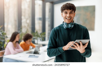 Happy young man with stylish headphones around his neck holds digital tablet, engaging with technology while his classmates work in the backdrop - Powered by Shutterstock