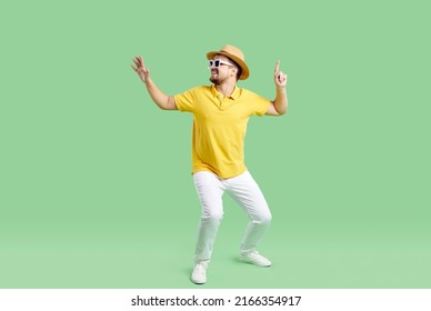 Happy Young Man In Summer Clothes, Hat And Sunglasses Dance On Green Studio Background. Overjoyed Male In Casualwear Have Fun Make Funny Dancer Moves. Entrainment And Party.