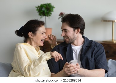 Happy young man teaching laughing mature senior mother using cellphone applications, watching funny photo video content online in social network, shopping in internet store, modern tech addiction.