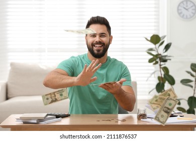 Happy young man throwing money at wooden table indoors