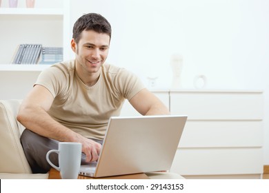 Happy Young Man In T-shirt Sitting On Sofa At Home, Working On Laptop Computer, Smiling.