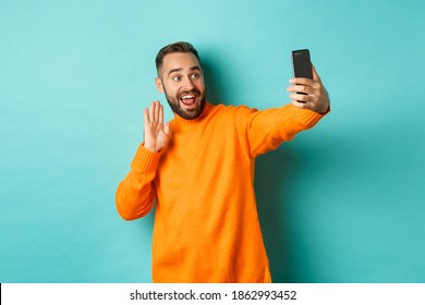 Happy Young Man Video Calling, Talking Online With Mobile Phone, Saying Hello To Smartphone Camera And Waving Hand Friendly, Standing Over Light Blue Background