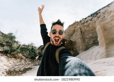 Happy Young Man Wearing Backpack Hiking On A Mountain Taking A Selfie With Smartphone And Action Cam.