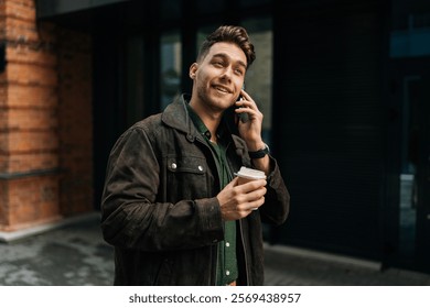 Happy young man wearing casual attire, holding takeaway coffee cup, engaging in phone conversation while strolling outside modern building in urban setting. Concept of successful city lifestyle. - Powered by Shutterstock