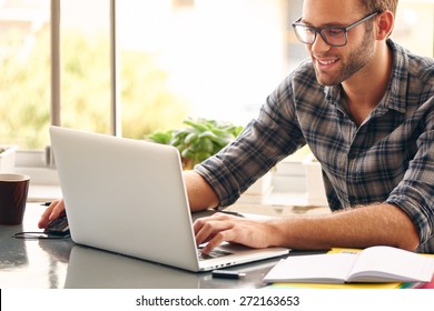 Happy young man, wearing glasses and smiling, as he works on his laptop to get all his business done early in the morning with his cup of coffee - Powered by Shutterstock