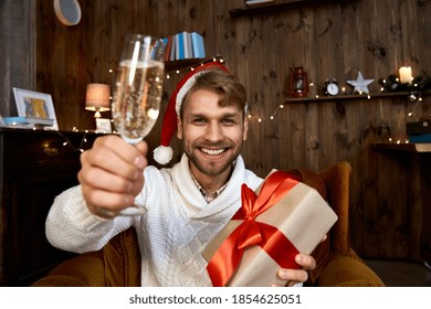 Happy Young Man Wearing Santa Hat Holding Christmas Gift Looking At Camera. Excited Guy Drinking Champagne Having Virtual Chat With Friend Celebrating New Year Distance Online Party. Web Cam View