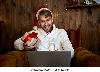 Happy Young Man Wears Santa Hat Saying Toast Celebrating Virtual New Year Party Using Laptop Holding Champagne Glass And Christmas Gift Having Distance Online Remote Chat Meeting With Family At Home.