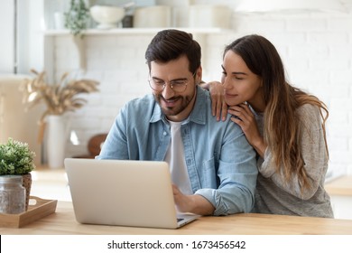 Happy young man and woman hugging, using laptop together, looking at screen, loving couple shopping or chatting online, using internet banking services, reading news in social network
