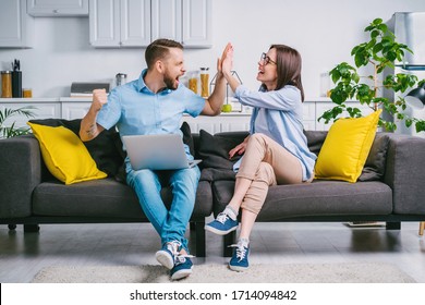 Happy Young Man And Woman Sitting At Home On Comfortable Sofa With Their Laptop Computer, Celebrating Money Win In Online Lottery Giving High Five To Each Other. 