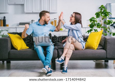Happy young man and woman sitting at home on comfortable sofa with their laptop computer, celebrating money win in online lottery giving high five to each other. 