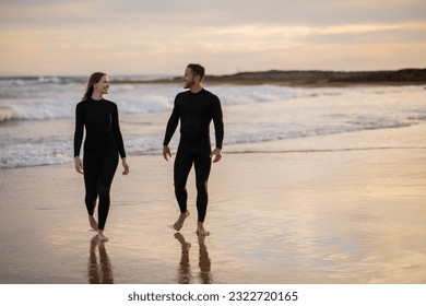 Happy young man and woman in wetsuits walking on the beach at sunset time, romantic millennial couple chilling at seashore, going out of water after surfing together, full length, copy space - Powered by Shutterstock