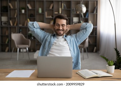 Happy young man worker in eyeglasses relaxing on chair with folded hands behind head, enjoying break pause time during working day or finishing online project on computer in modern home office.