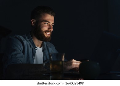 Happy Young Man Working On Laptop Computer While Sitting At The Table Indoors At Night