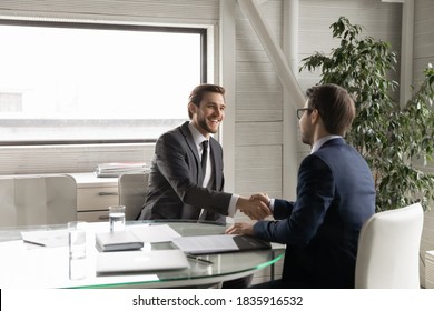 Happy young manager shaking hands with leader, feeling thankful for new job offer. Satisfied with good working results ceo executive manager praising male employee at meeting, sitting at table.