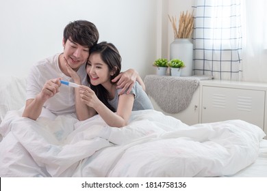 Happy Young Married Asian Couple Looking At Pregnancy Test Together On Bed In White Bedroom.