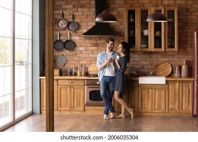Happy young married couple enjoying morning coffee in kitchen of new apartment. Barefooted dating guy and girl standing together on heat floor at home, hugging, drinking hot beverage. Full length