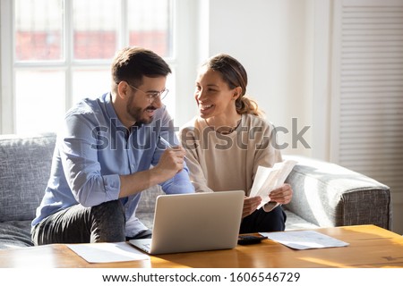 Happy young married couple sit on couch calculate expenses use easy online banking service at home, smiling millennial husband and wife count taxes house expenditures pay bills on internet