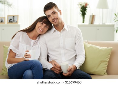 Happy Young Married Couple Sitting On The Sofa With Cups Of Tea