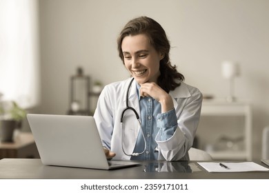 Happy young medical practitioner woman working at laptop, typing, smiling, laughing, sitting at table, using Internet technology for online communication, chatting with patients - Powered by Shutterstock