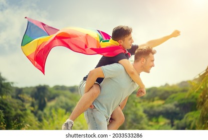 Happy Young Men, Gay Family With Flying LGBT Rainbow Flag Having Fun Outdoors. Joyful Gay Couple Together In A Park. Laughing, Hugging, Enjoying Nature Outside, Over Sunny Sky. Super Hero. Piggyback