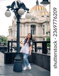 Happy young mexican woman in embroidered top looking at camera while standing in street and touching lamppost in front of blurred Bellas Fine Arts Palace in Mexico City in daylight