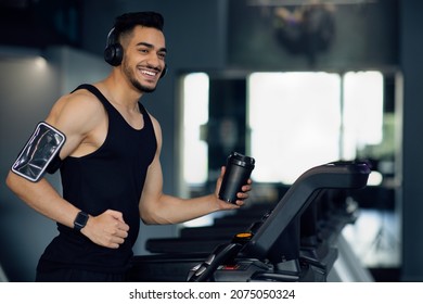 Happy Young Middle Eastern Male Athlete Jogging On Treadmill At Gym, Cheerful Muscular Arab Man Holding Sport Shaker Bottle And Wearing Wireless Headphones, Listening Music While Training, Copy Space - Powered by Shutterstock