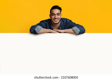 Happy young middle eastern man in casual outfit leaning on huge horizontal empty placard for advertisement over yellow studio background, cheerfully smiling at camera, recommending something nice - Shutterstock ID 2107608005