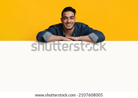 Happy young middle eastern man in casual outfit leaning on huge horizontal empty placard for advertisement over yellow studio background, cheerfully smiling at camera, recommending something nice
