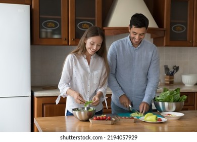 Happy Young Millennial Couple Cooking Fresh Salad For Homemade Dinner Together, Slicing Fresh Vegetables Into Metal Bowl On Kitchen Table, Eating Natural Organic Food, Keeping Healthy Diet