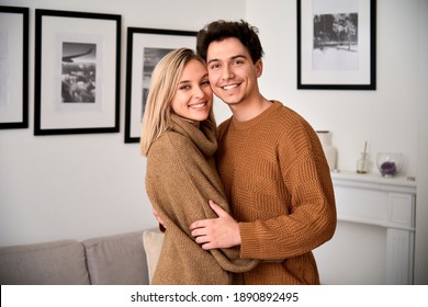 Happy Young Millennial Couple Hugging Looking At Camera Standing At Home. New Modern Apartment Owners, Real Estate Buyers Embracing Posing In Living Room, Portrait. Mortgage Investment Concept.