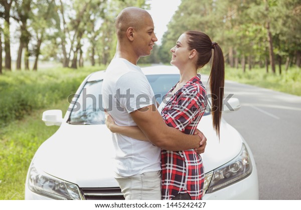 Happy young mixed couple\
hugging on a country road, while travelling by car. Beautiful\
loving couple enjoying their roadtrip, taking break from driving,\
admiring nature