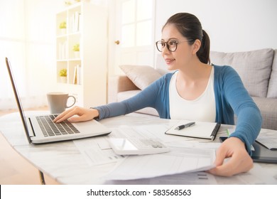 Happy Young Mixed Race Asian Chinese Woman Calculating Bills By Laptop Computer Sitting On Sofa In The Living Room At Home. Interior And Domestic Housework Concept.