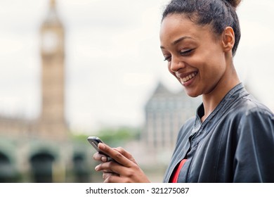 Happy young mixed race businesswoman portrait outdoors in London with Big Ben as background while using smartphone to send a text message. Filtered image.