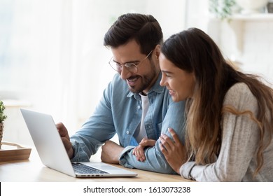 Happy young mixed race couple enjoying free weekend time, watching funny videos on computer. Smiling woman embracing husband, looking at laptop screen, shopping, web surfing together online.