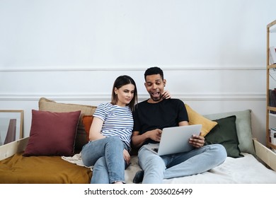 Happy Young Mixed Race Couple Enjoying Time At Home Using Computer Sitting On Sofa, Black Man And White Woman Having Fun Online Shopping Or Surfing Web Relaxing On Couch