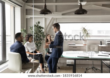 Happy young mixed race teammates enjoying break pause time with leader, having funny talk. Smiling multiracial employees involved in workshop with skilled 30s male business trainer mentor in office.