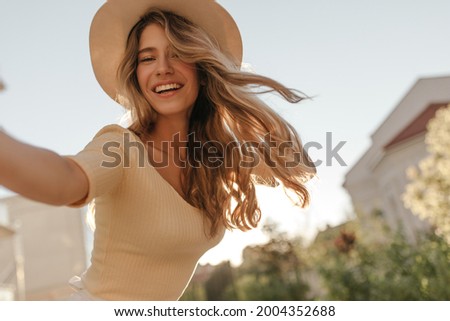 Happy young model makes photo holding camera in hand close-up. Her hair is flying in wind, she is wearing hat. Outdoor photo.