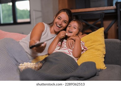 Happy young mom and child girl laughing holding snack popcorn remote control enjoy funny television comedy movie