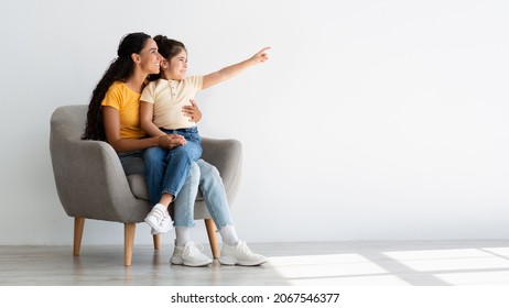 Happy Young Mom And Cute Female Child Pointing Aside While Relaxing In Armchair At Home, Cheerful Middle Eastern Mother And Daughter Showing Copy Space For Young Design Or Advertisement, Panorama - Powered by Shutterstock