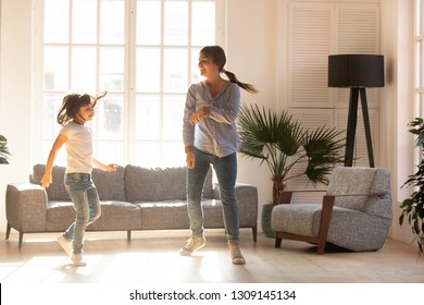 Happy Young Mom And Cute Little Daughter Jumping Dancing In Modern House Living Room, Joyful Mother With Child Girl Having Fun Enjoy Moving To Music Together At Home, Mum And Kid Funny Activity