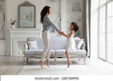 Happy young mom and cute little preschooler daughter have fun dancing jumping in living room together, overjoyed millennial mother or nanny feel playful entertain at home with small girl child