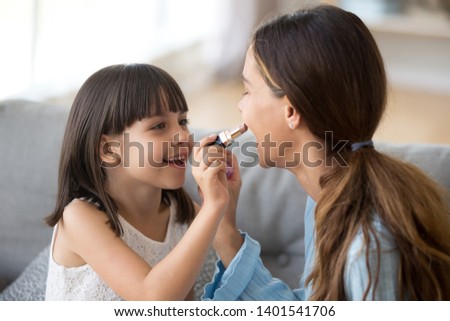 Happy young mom and cute preschooler daughter have fun together at home paint lips with lipstick, excited nanny or sister entertain do makeup with small girl child, enjoy spend time at home
