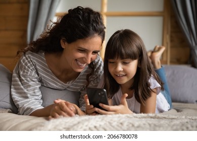 Happy Young Mom And Daughter Kid Relaxing On Bed, Holding Smartphone, Using Online App, Virtual Service, Playing Game, Making Call, Reading Book, Watching Media Content, Shopping On Internet