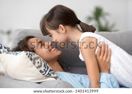 Happy young mom have fun playing with funny little daughter, relax on couch touching noses, smiling mother rest enjoy spending time with preschooler kid having tender sweet moment at home