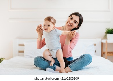 Happy Young Mom Helping To Her Infant Child Making First Steps, Sitting On Bed At Home And Smiling At Camera, Spending Time With Little Kid, Copy Space