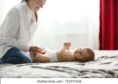 Happy Young Mom In Light Bedroom Smiles And Dressing Her Precious Baby Boy For A Morning Walk In Park. Family Concept..